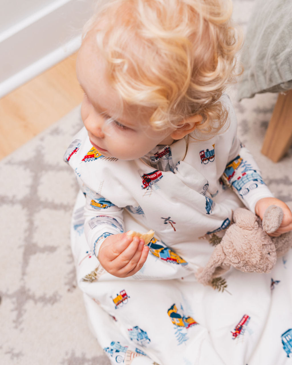 Child wearing a white onesie with a colorful train pattern, sitting on a wooden floor.