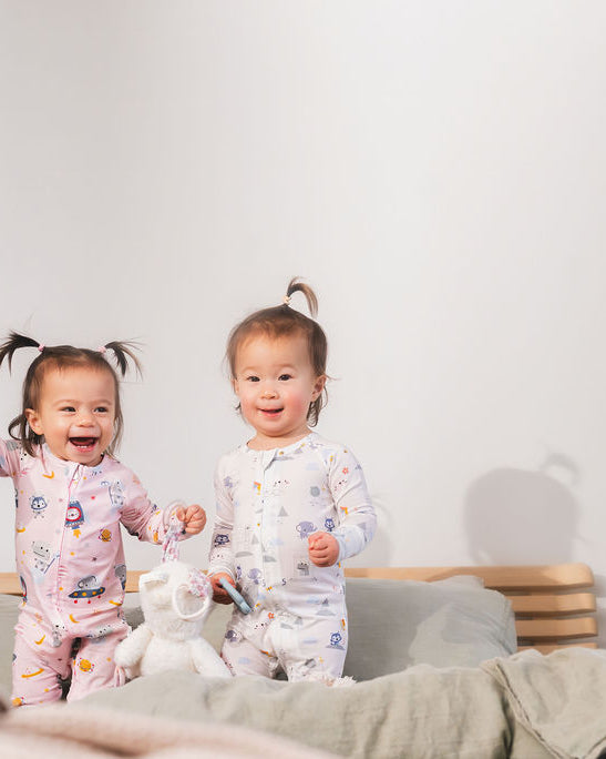 Two children in pajamas standing on a bed with a plush toy and a stuffed animal.