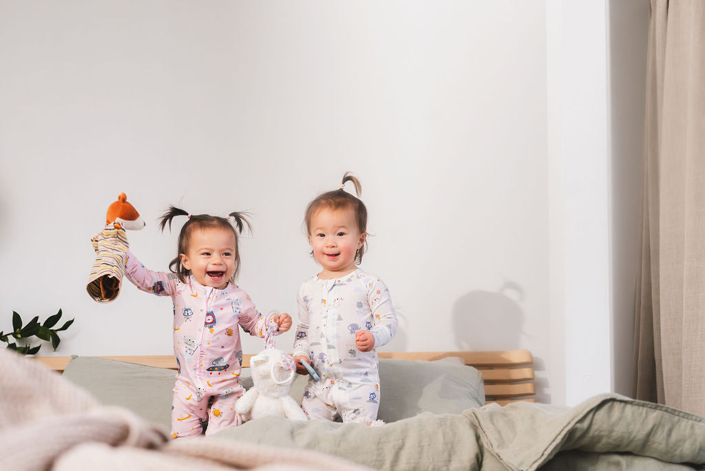Two children in pajamas standing on a bed with a plush toy and a stuffed animal.